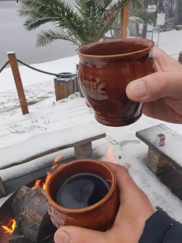 Dwie osoby trzymają glinianych kubków z gorącym naparem przy ognisku na plaży zimą, w tle jezioro i palmy.