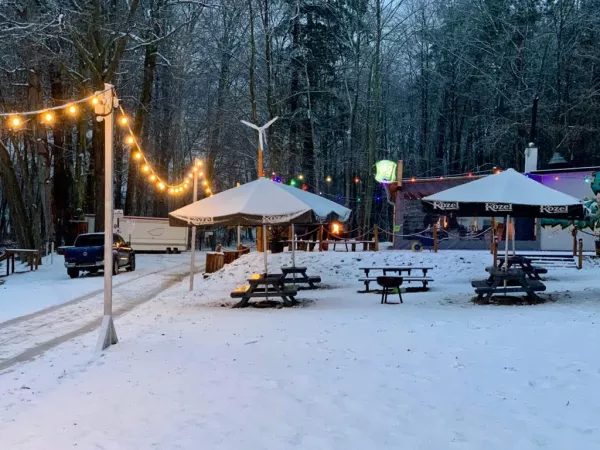 Zimowa plaża z drewnianymi stołami do ogniska, białymi namiotami, oświetleniem i restauracją Kozela w tle.