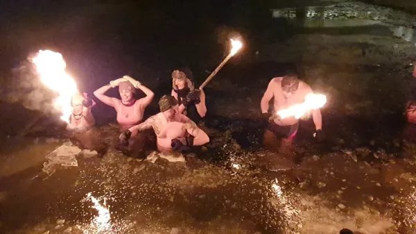 Grupa ludzi w wodzie nocą przy plaży, trzymająca płonące pochodnie, podczas zabawy nad jeziorem.