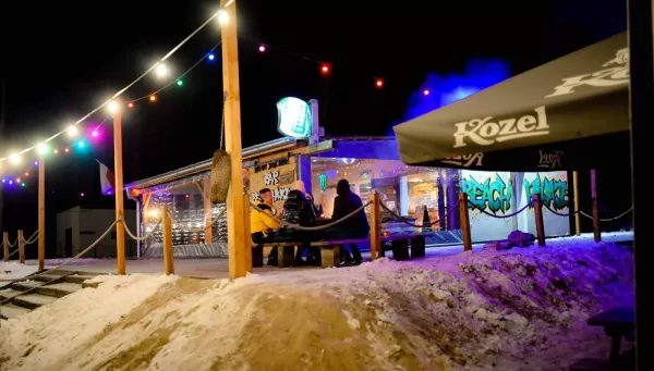 Nocny widok na plażę z drewnianą restauracją oświetloną kolorowymi lampkami, gdzie kilka osób siedzi na ławce, a w tle widać zaśnieżony teren ośrodka.