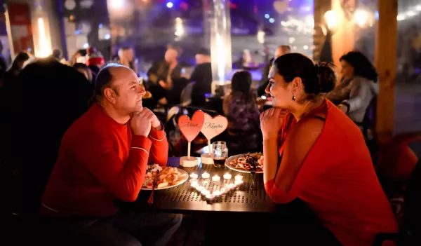 Dwie osoby w czerwonych ubraniach siedzą przy stole z jedzeniem i dekoracją sercami, w tle widać inne gości i oświetlenie restauracji.
