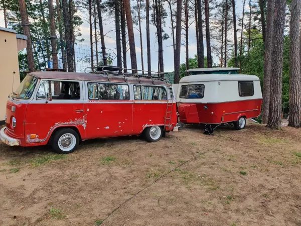 Czerwony zabytkowy autobus i przyczepa kempingowa zaparkowane wśród sosen na terenie ośrodka.