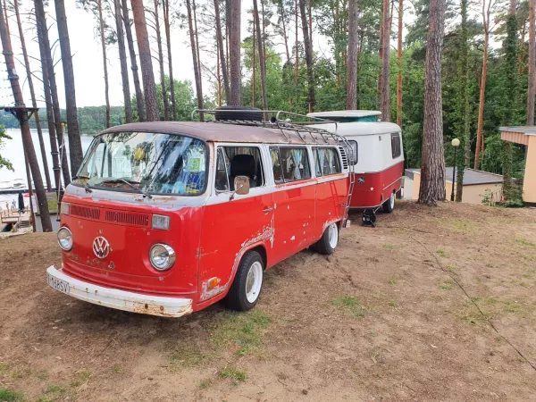 Czerwony zabytkowy autobus Volkswagen i przyczepa kempingowa zaparkowane między sosnami nad jeziorem.