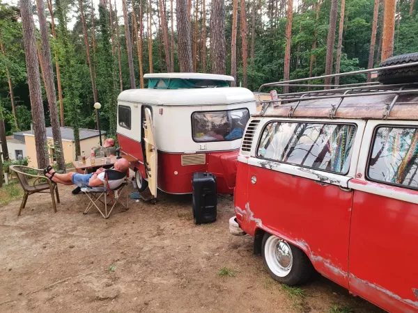 Camping w lesie z zabytkową przyczepą kempingową i samochodem dostawczym, osoba relaksuje się na krześle.